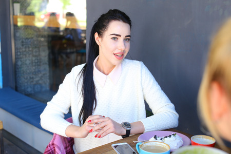 Beautiful sweet young lady girl smiles, chatting and gossiping with friends over cup of coffee at table at wooden table on gray background panoramic windows and gray walls. Girl with black hair dressed in peach shirt and white knitted sweater.Concept of mの写真素材