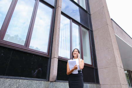 Serious young adult business woman  talks on phone, solves problems, holds and looks at documents and stands on background of gray wall of business center. Woman with black hair, dressed in white blouse with black skirt and heels. Concept of business meetの写真素材