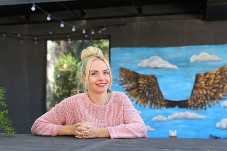 Lovely young adult female woman smiles broadly, looks and posing at camera, sits at wooden table on gray background panoramic windows, walls decorated with pictures and garland of lanterns. Girl with blond hair dressed in pink knitted sweater. Concept of の写真素材