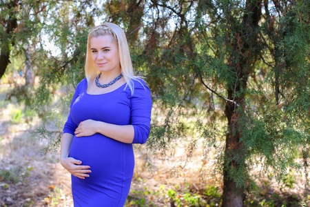 Marvelous Young Adult Female to Carry Child Under Ones Heart, Relax and Enjoy Nature and Fresh Air, and Stands in Big Green Park Outdoors in Daytime. Girl With Blond Hair, Braided in Braid in Front Dressed in Long Blue Dress. Concept of Happy Pregnancy anの写真素材