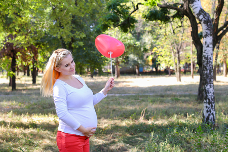 Alluring Future Mother of European Appearance Enjoys Pleasant Things and Look Forward to Appearance of First-Born Into World Photographed For Advertising of Womens Clothing For Pregnant With Pink Ballon in Shape of Heart in Green Sunny Park Outdoors in Daの写真素材