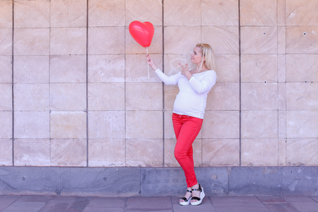 Beautiful Female of European Appearance to Carry Child Under Ones Heart, Holding in Hand Pink Balloon in Shape of Heart, Smiling and Posing at Camera in full growth, Raises Thumb up and Stands on Background of Stone Slabs in Open Air in Daytime. Girl Withの写真素材