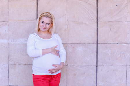 Attractive Young Adult Female to Carry Child Under Ones Heart, Looking in Camera and Tummy, Happy Posing For Family Album and Stands on Background of Stone Slabs in Open Air in Daytime. Girl With Blond Hair, Braided in Braid in Front Dressed in White Blouの写真素材