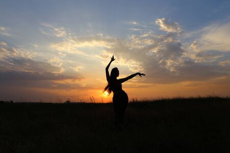 Female with beautiful figure takes yoga postures, gymnastics and ballet spends stretching and meditating, posing for advertising shoot fitness studio, healthy energy drink, dancing belly dancing among wide field on background of yellow sunset and beautifuの写真素材