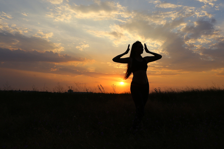 Female with beautiful figure takes yoga postures, gymnastics and ballet spends stretching and meditating, posing for advertising shoot fitness studio, healthy energy drink, dancing belly dancing among wide field on background of yellow sunset and beautifuの写真素材