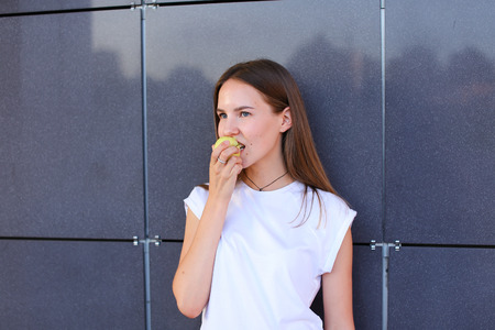 Young beautiful girl holds in hand and bites small green apple against background of gray wall. Girl with long light brown hair dressed in white shirt. Concept of Fruit, healthy eating, lunch break, green vitamin.の写真素材