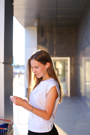 Business attractive girl young business woman holds white telephone, listens to music, uses phone to work, solves problems, smiles and stands in profile on background of gray wall of business center. Girl with long light brown hair dressed in white shirt の写真素材