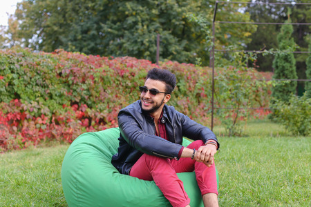 Arabian Young Man Sitting and Resting From City Bustle in Soft Green Chair, smiles and Looks Around. Handsome Guy Has Dark Hair and Black Beard and dressed in Maroon Shirt Over Black Leather Jacket and Bright Pink Jeans and White Sneakers Accessories Fromの写真素材
