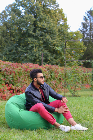 Arabian Young Man Sitting and Resting From City Bustle in Soft Green Chair, smiles and Looks Around. Handsome Guy Has Dark Hair and Black Beard and dressed in Maroon Shirt Over Black Leather Jacket and Bright Pink Jeans and White Sneakers Accessories Fromの写真素材