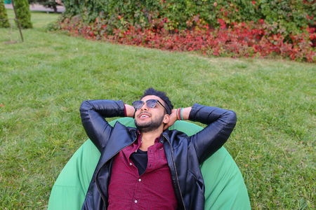 Portrait of Young Arab Masculine Man Student, Which Lies on Soft Green Chair, Dreams, Meditate, Thinking About Something, Smiling and Looking at Heaven, Hands Behind Head on Background of Green Grass Outdoors. Stylish Guy With Dark Hair Dressed in Black Tの写真素材