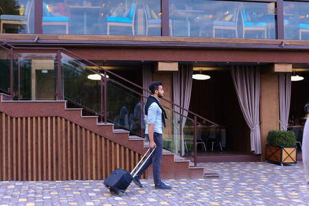Arabic Muslim Young Man Coming Out of Corner and Goes With Travel Bag to Modern Restaurant, Stopped and Smiling, Holding Black Suitcase. Handsome Muscular Guy Dressed in Black Classic Trousers, White Shirt on Top of Which Knotted Black Sweater. Concept ofの写真素材