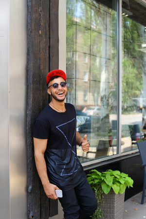 Handsome young man arab stands near wall smiles, rejoices and holds in hand mobile phone on background of panoramic windows in modern restaurant. Man dressed in dark T-shirt, black pants, white sneakers and black red color cap and sunglasses. Concept of lの写真素材