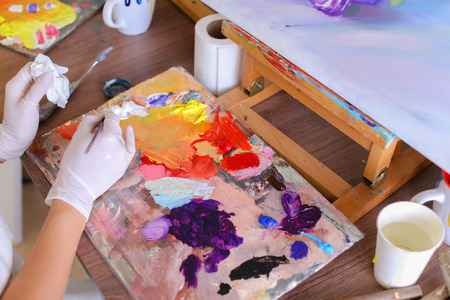 Women Artist extrudes hands in white gloves oil paint from tubes on multi-colored palette for viewing and mixing colors to start drawing pictures in art studio. On table stands glass of water, jar of colored pencils and brushes and palette sketch. Conceptの写真素材