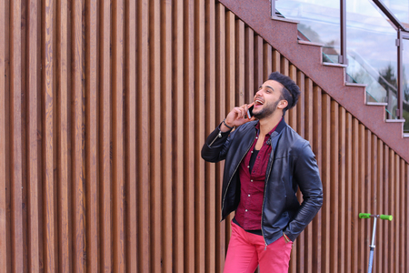 Confident Attractive Young Guy Muslim Businessman Goes Slowly Along Wall, Holding Phone in Hand and Talks, Resolve Issues, Discuss Future Plans and Smiling, Holds Hand in Pocket, Stands on Background of Wooden Panel Stairs Near House Outdoors. Bearded Manの写真素材