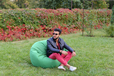 Arabian Young Man Sitting and Resting From City Bustle in Soft Green Chair and Looks Around. Handsome Guy Has Dark Hair and Black Beard and Wearing Maroon Shirt Over Black Leather Jacket and Bright Pink Jeans and White Sneakers Accessories From Sunglassesの写真素材