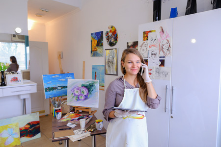 Female European appearance painter talking on mobile with clients, solve problems, appointments with students and posing for advertising art appliances or art school, order paint, canvas or frame.Woman With Long Light Brown Hair Dressed Sht Pale Purple blの写真素材