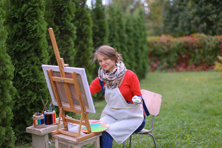 Cute female painter posing, laughing and in front of camera , draws and smiling with brushes and colorful palette in hand, sits on chair near easel in green park outdoor.  Girl with brown hair dressed in red jamper, dark denim overalls and white apron, chの写真素材