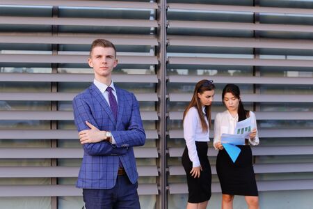Handsome confident young male guy businessman, office worker, student posing in front for camera,crossed arms in front of on chest, posing for shoot of modern mens business clothing catalog and stands at colleagues background two attractive young women, bの写真素材