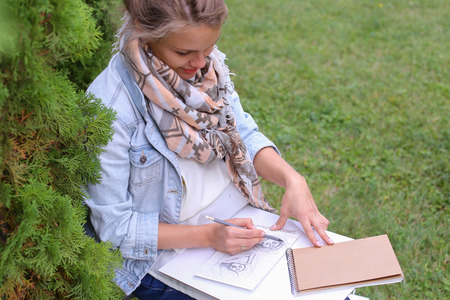 Contemporary Artist Works on Facial Contour of Depicted on Portrait Unknown Girl, Draws Sketch of Fictional Head, Prepares Gift For Friend. Woman Located in Park Outdoors in Background Stands Bench With Colors and Additional Art Materials. Artist Dressed の写真素材