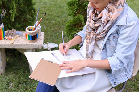 Contemporary Artist Works on Facial Contour of Depicted on Portrait Unknown Girl, Draws Sketch of Fictional Head, Prepares Gift For Friend. Woman Located in Park Outdoors in Background Stands Bench With Colors and Additional Art Materials. Artist Dressed の写真素材