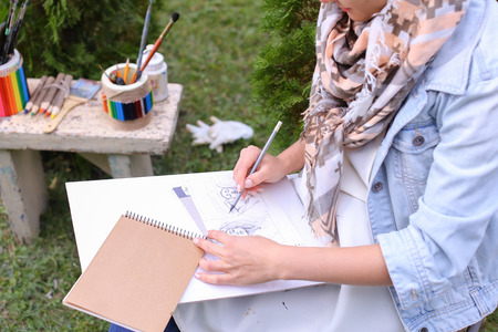 Contemporary Artist Works on Facial Contour of Depicted on Portrait Unknown Girl, Draws Sketch of Fictional Head, Prepares Gift For Friend. Woman Located in Park Outdoors in Background Stands Bench With Colors and Additional Art Materials. Artist Dressed の写真素材