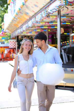 Beautiful young people, loving couple strolling in amusement park, looking at each other, kissing and cuddling, try candy floss, pose for camera and fooled at an amusement park outdoors. Girl with long blond hair and sunglasses dressed in white top and beの写真素材