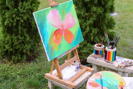 Photo completed and unfinished picture, works of art on which  image multi-colored butterfly on canvas and stands on wooden easel, palette and brush lying near easel, on table stands colorful banks in which there are other artistic intrumenty and colored の写真素材