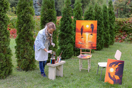 Artist cleans workplace, evaluate work that stands on easel, smiling, posing and looking in camera, standing in full length near paintings in park outdoors on background of coniferous plants in afternoon. Girl dressed in red sweater, dark jeans, white aprの写真素材
