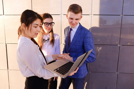 Modern young man and two attractive smart young women, business people, students communicate, holding laptop and study work plan, familiarize with presentation, teach each other new programs, solve important problems and workloads holding folder with docuの写真素材