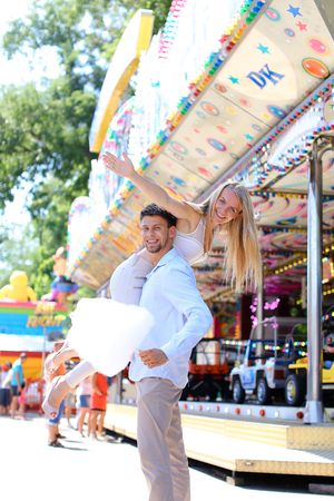 Sweethearts cuddling, pose for camera and fooled, boy holding girl with hand on shoulder at an amusement park outdoors. Girl with long blond hair dressed in white top and beige pants, with small blue bag on shoulder, guy dressed in blue light shirt and beの写真素材
