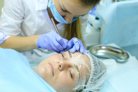 Young blonde woman professional, engaged in reconstruction work responsibly, eyebrows manual method performs procedure, using thinnest needle and maniples, draws most natural eyebrow, side by side on couch and ready for client procedure in cosmetic white の写真素材