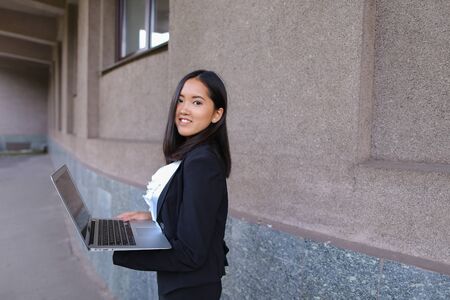 Beautiful young female woman asian girl entrepreneur successful person holds laptop in hands and works, solves problems, and stands on background wall of  business buildings .Woman with black hair, dressed in white blouse and classic jacket with black skiの写真素材