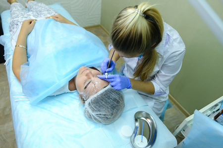 Young blonde woman professional, engaged in reconstruction work responsibly, eyebrows manual method performs procedure, using thinnest needle and maniples, draws most natural eyebrow, side by side on couch and ready for client procedure in cosmetic white の写真素材