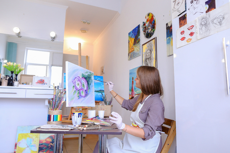 Charming Young Woman Artist Draws Oil Painting With Purple Flowers on a White Canvas, Female Enjoying Favorite Work, Hobbies, posing  for camera and Painting Picture to Order or Gift. Woman With Long Light Brown Hair Dressed Sht Pale Purple Jacket, White の写真素材
