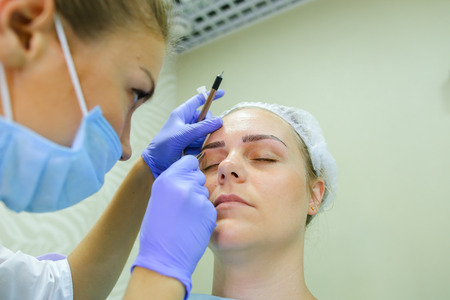 Successful female browmaker consult client girl completes registration and drawing eyebrow hairs and antiseptic wipes. Master dressed in white robe, blue rubber gloves and face protection gauze bandage. Concept of mikrobleyding eyebrows micropigmentation の写真素材