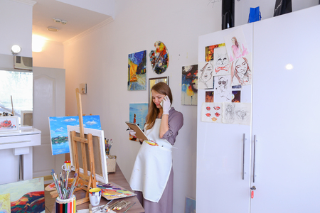 Lovely Female Painter Orders Things to Draw or Picture Frames , Clarifies Meeting With Students For Master Class, Communicates With Clients About Sale of Paintings. Woman With Long Light Brown Hair Dressed Sht Pale Purple Jacket, White Apron and White Gloの写真素材