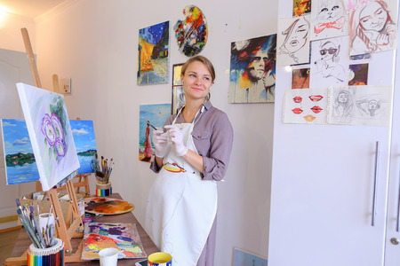 Artist attractive woman of European appearance writes oil painting, enjoying things what love , paint picture for sales and order, listening to music on headphones through phone, stands and works in bright art studio against background of wall hung with pの写真素材