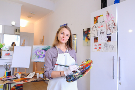 Cute female painter posing, laughing and fooling around in front of camera and smiling with brushes and colorful palette in hand, standing in studio with white art paintings on walls. Girl European appearance advertises art school for adults and children,の写真素材