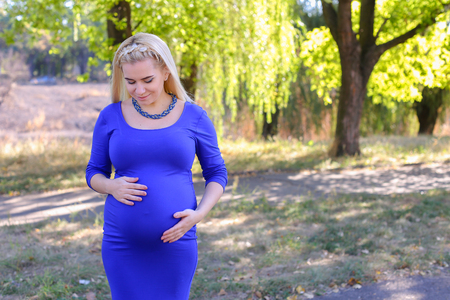 Beautiful Young Woman in Expectation Baby Goes Towards Husband, to Meet in Cafe, Going to Spend Time With Friends or go to Visit Parents, Rest From Urban Bustle of Breathing Fresh Air in Park Outdoors in Daytime. Blonde With Hair Braided in Pigtail in Froの写真素材
