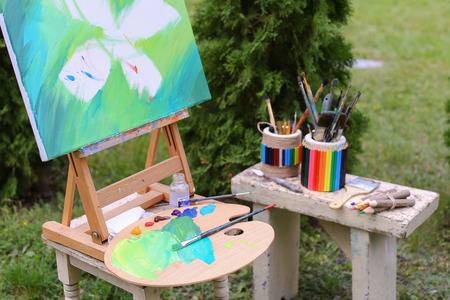 Photo completed and unfinished picture, works of art on which  image multi-colored butterfly on canvas and stands on wooden easel, palette and brush lying near easel, on table stands colorful banks in which there are other artistic intrumenty and colored の写真素材