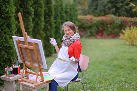 Cute female painter posing, laughing and in front of camera , draws and smiling with brushes and colorful palette in hand, sits on chair near easel in green park outdoor.  Girl with brown hair dressed in red jamper, dark denim overalls and white apron, chの写真素材