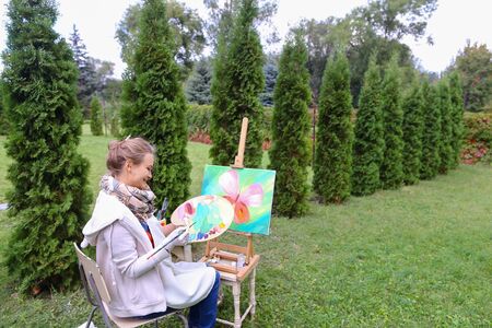 Lovely girl artist paints picture, presses  paint and mix , relaxes and relieves stress engaging in favorite hobby, evaluating look at  picture, advertise art school. Girl with brown hair collected in beam dressed in light jacket, red jacket, dark denim oの写真素材