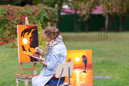 Young cute woman artist  finishes painting which depicts woman who keeps  sun on orange background, sitting with his back to  photographer and  behind wooden easel, holds palette smeared in paint in large park green day outdoors.Girl dressed in red sweateの写真素材