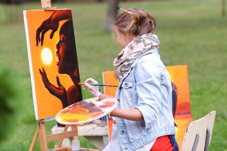Young cute woman artist  finishes painting which depicts woman who keeps  sun on orange background, sitting with his back to  photographer and  behind wooden easel, holds palette smeared in paint in large park green day outdoors.Girl dressed in red sweateの写真素材