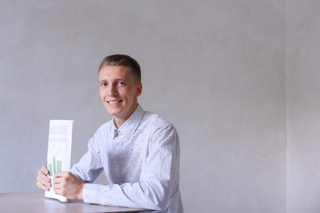 Entrepreneur looking into camera and smiling, completes work day. Men with short hair and blond hair sitting on chair at white table in office and dressed in white classic shirt. Concept of businessman office work, sedentary routine work documentation, suの写真素材