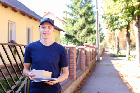 Young Courier European Appearance Like Job Carry Parcel. Guy in Black T-Shirt and Cap Bears Welcome Box For Customers, Delivery Man Work With Pleasure Reaches His Hand and Gives Box With Order. Male Good Mood on Warm Sunny Day, Looking at Camera and Smiliの写真素材
