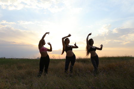 Three beautiful girls dance and show movements of Eastern dance, standing in unusual posture and meditate at sunset, shot to advertise fitness club or dance studio in open air in broad field against sky and sunset. Girl with shoulder-length hair dressed iの写真素材