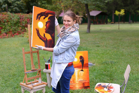 Female European appearance artist evaluates picture and removes from easel, holds and posing with picture which depicts woman with sun in hands made orange smiling and standing in full growth on background of lawn and angered by conifers in large park in の写真素材