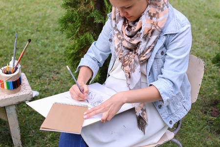 Contemporary Artist Works on Facial Contour of Depicted on Portrait Unknown Girl, Draws Sketch of Fictional Head, Prepares Gift For Friend. Woman Located in Park Outdoors in Background Stands Bench With Colors and Additional Art Materials. Artist Dressed の写真素材