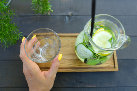 Glass decanter with refreshing drink from cucumber, lemon, mint and empty glass faceted cup which is held in the hand stands on wooden board on gray wooden table on background of green flowers in gray bucket, iron pot in cafe. Concept of refreshing drink,の写真素材
